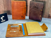 May include: Three personalized leather journals; one brown leather journal with the initials &#39;WA&#39; engraved, one orange leather journal with the inscription &#39;Never forget that you are loved&#39;, and one dark-purple leather journal with the name &#39;Olivia&#39; and a leaf design engraved.  All journals feature multiple pockets and card slots, and one includes a pen holder. The journals are displayed on a wooden surface.