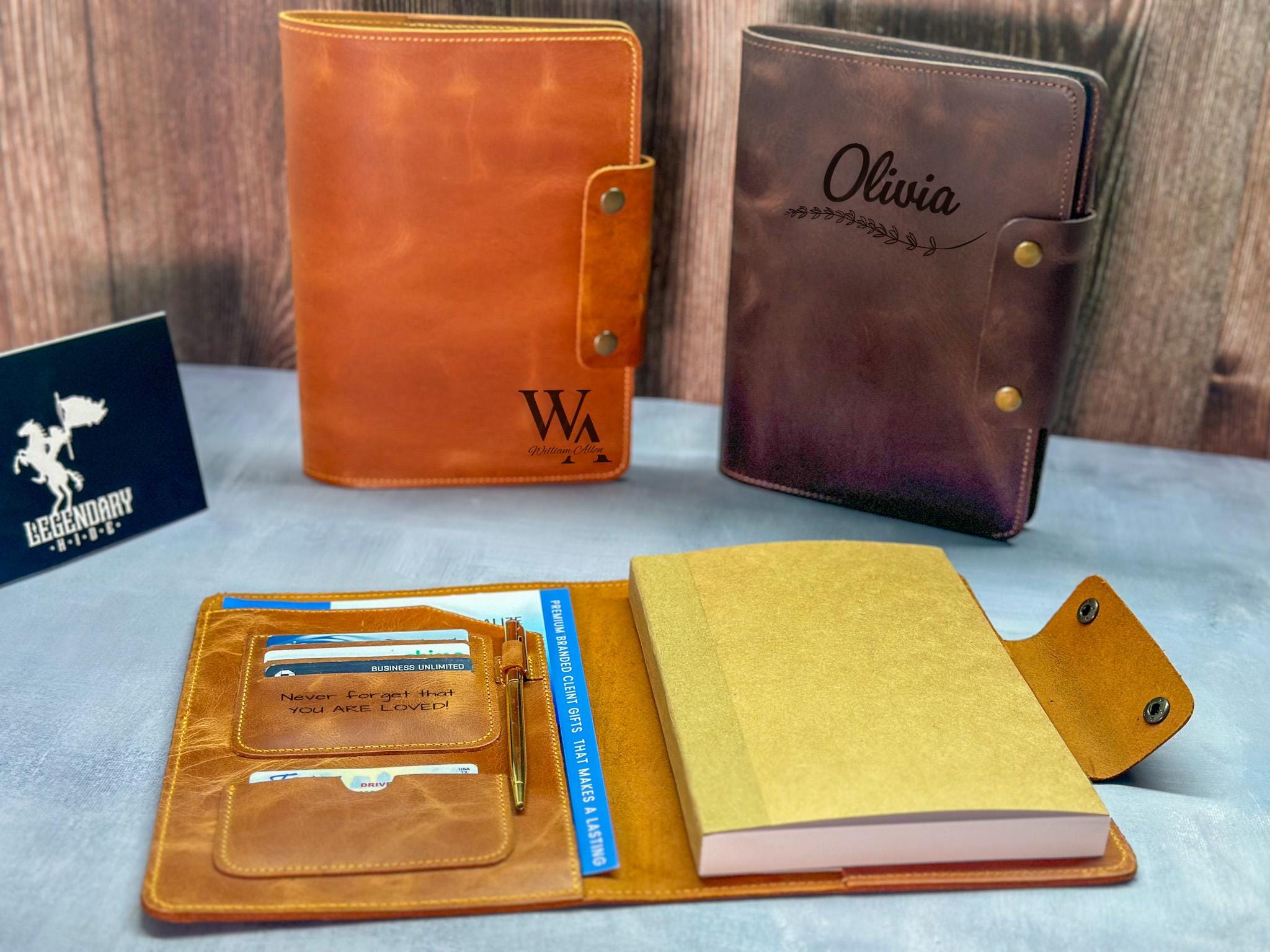 May include: Three personalized leather journals; one brown leather journal with the initials &#39;WA&#39; engraved, one orange leather journal with the inscription &#39;Never forget that you are loved&#39;, and one dark-purple leather journal with the name &#39;Olivia&#39; and a leaf design engraved.  All journals feature multiple pockets and card slots, and one includes a pen holder. The journals are displayed on a wooden surface.